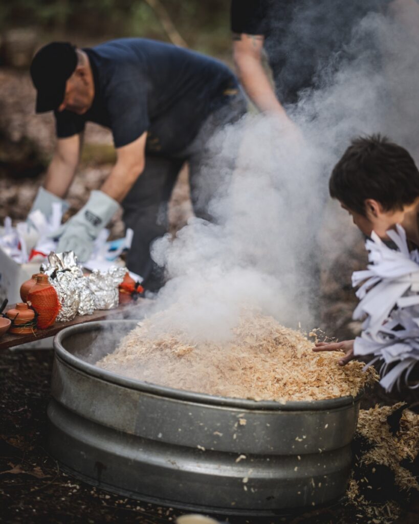 A person pouring wood shavings into a smoking galvanized metal fire pit while preparing an outdoor primitive pottery firing in the woods.