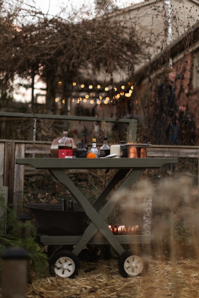 A rolling wooden bar cart in a garden at dusk, topped with cocktail fixings, bottles, and copper mugs for an outdoor autumn gathering.