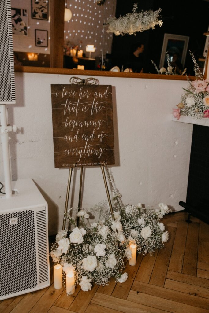A wooden wedding sign on a gold easel featuring an F. Scott Fitzgerald quote from The Great Gatsby.