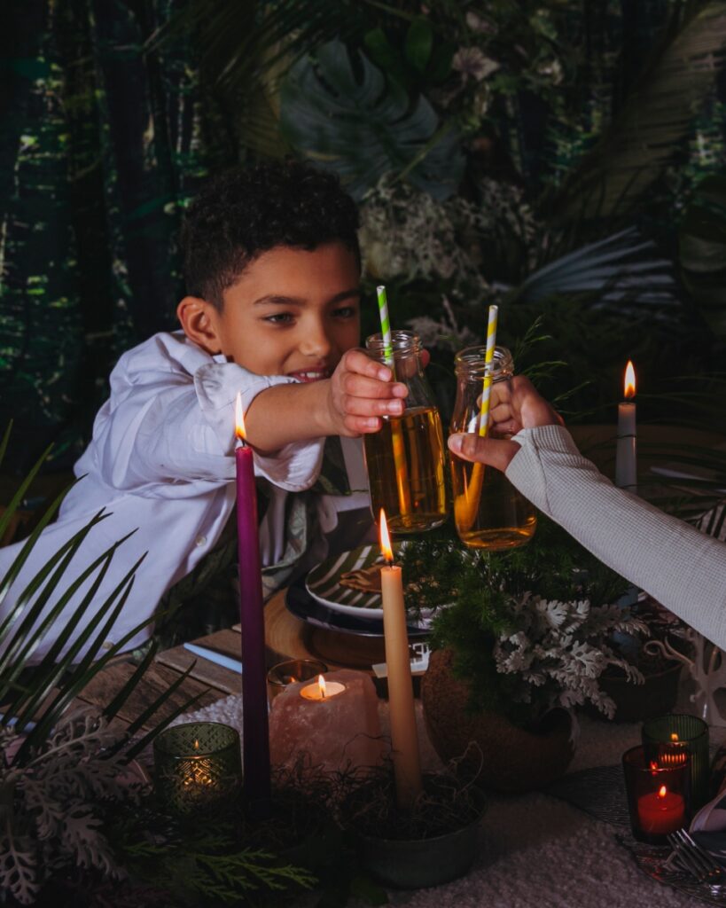 A young boy in a white button-down shirt toasts with a small glass milk bottle of apple juice and a striped straw at a candlelit forest-themed party.