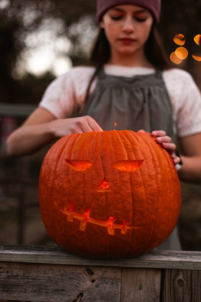 A young girl in a purple beanie finishing a carved jack-o'-lantern on a wooden railing during an outdoor fall celebration.