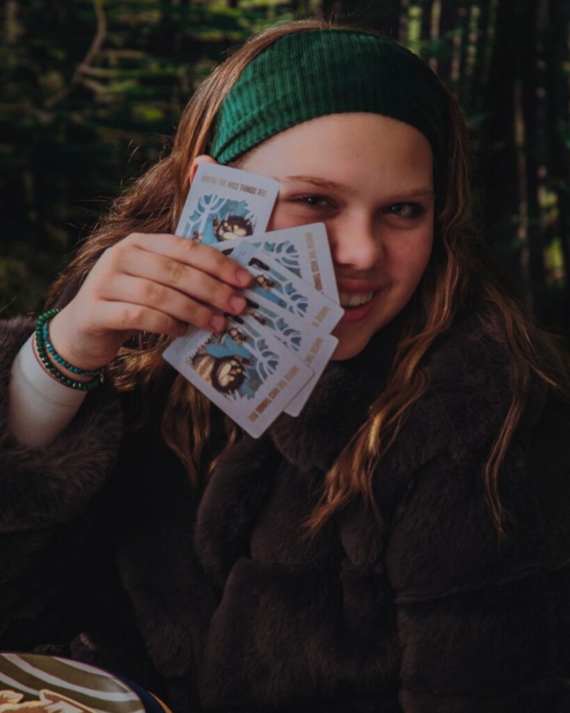A young girl in a brown faux fur coat and green headband smiling while holding a fan of "Where the Wild Things Are" themed playing cards.