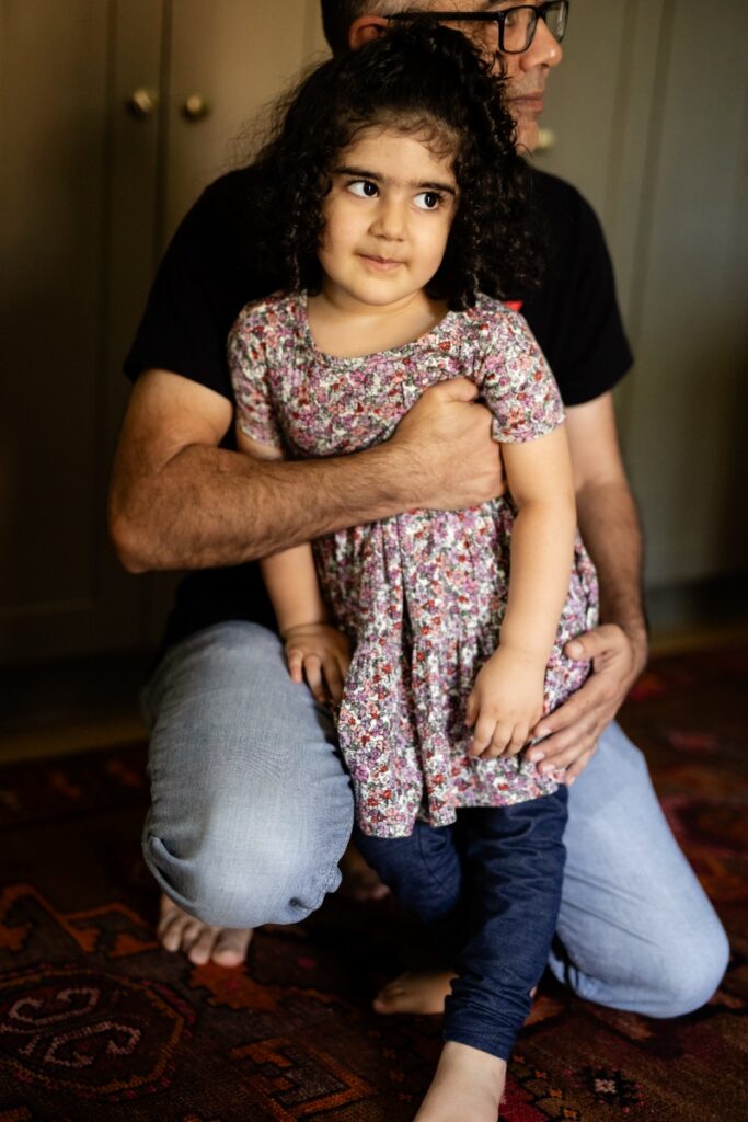A young girl with curly hair in a floral dress stands beside a man, both looking off-camera during an intimate gathering in a home.