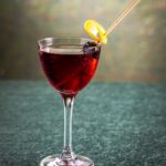 Artist in Residence cocktail in a stemmed glass, garnished with a Luxardo cherry wrapped in a lemon twist, featuring flavors of citrus gin, cherry, and chocolate bitters.