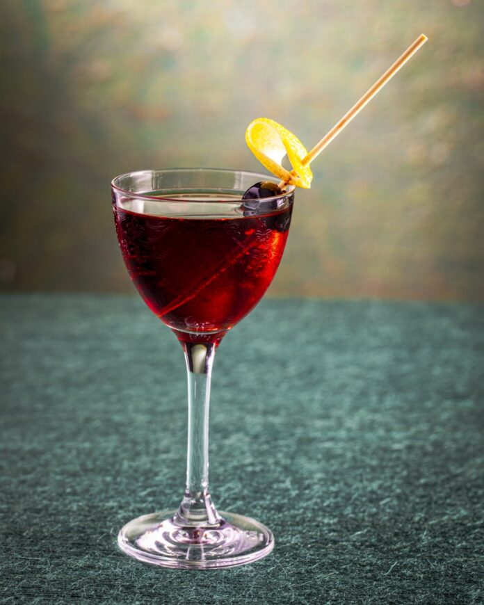 Artist in Residence Cocktail TABLE Magazine Artist in Residence cocktail in a stemmed glass, garnished with a Luxardo cherry wrapped in a lemon twist, featuring flavors of citrus gin, cherry, and chocolate bitters.