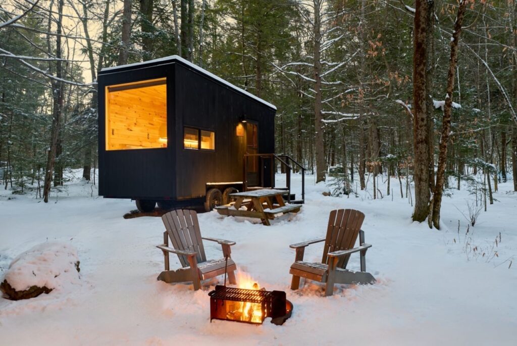 An outdoor black cabin with two chairs and a fire pit outside in the snow.