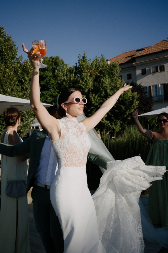 Bride Cristina Bartolacci, wearing a high-neck lace wedding gown and pink sunglasses, cheers with an orange cocktail during her outdoor reception.