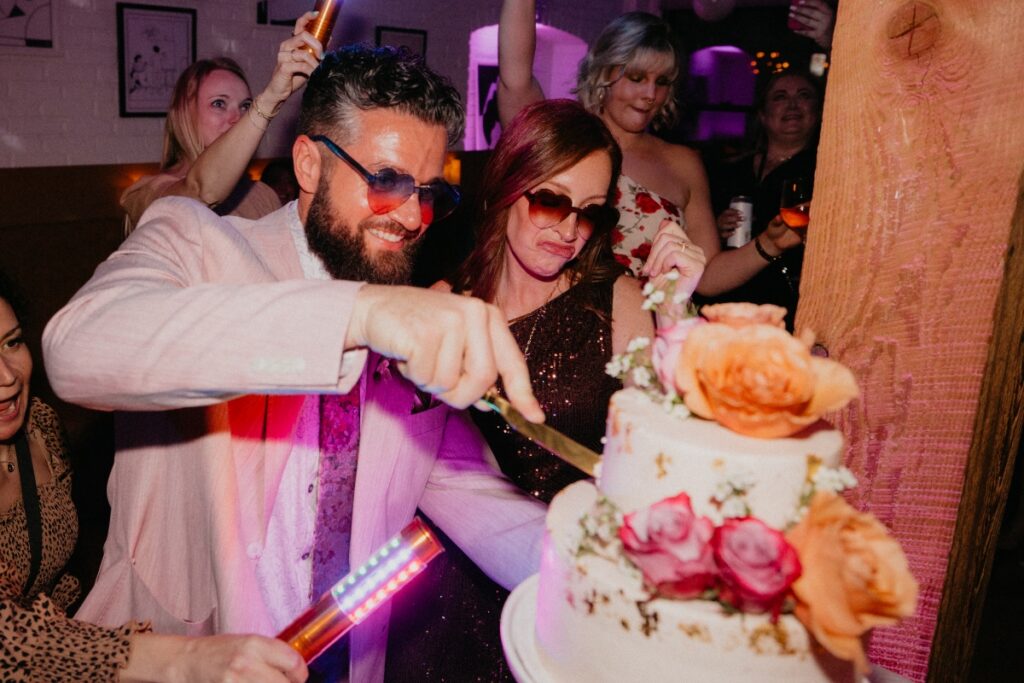 Groom Anthony and his wife cutting a tiered wedding cake adorned with orange and pink roses during their high-energy Pittsburgh reception.