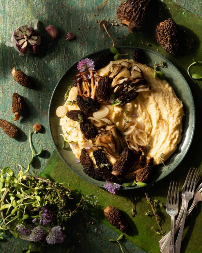 A green bowl on a green table holds cheesy grits inside mixed with small blossoms and springtime mushrooms with uncooked mushrooms all around the bowl.