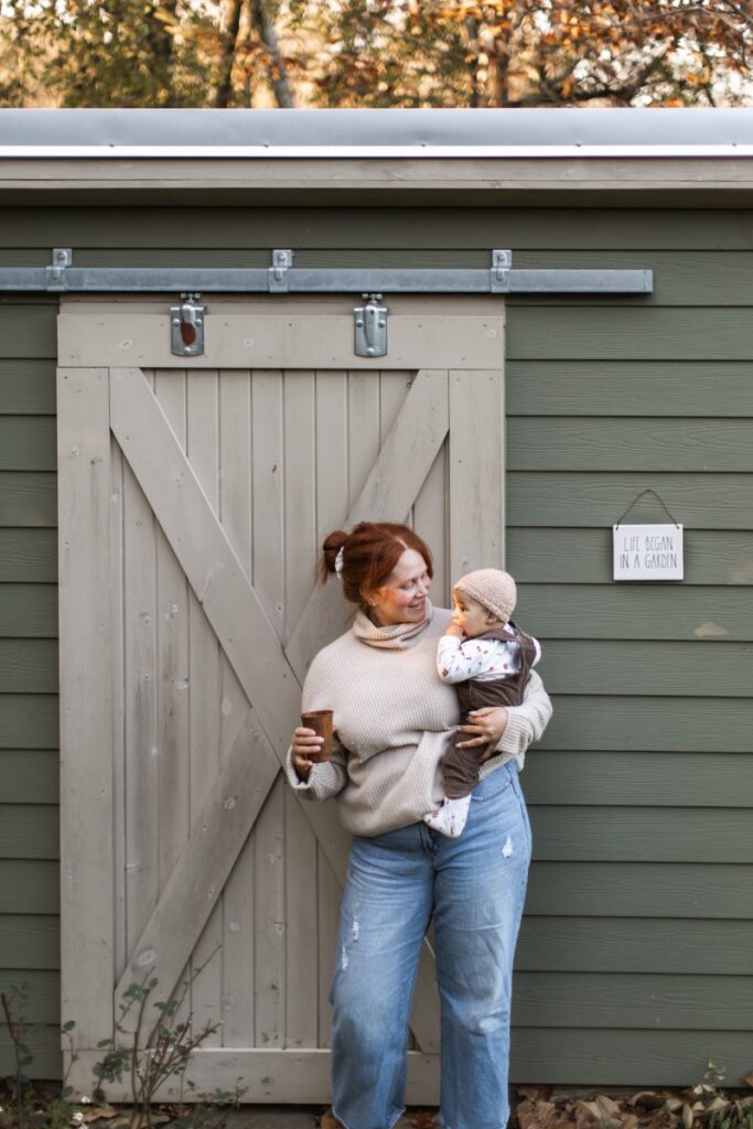A mother, Chelsea Barber, holding her baby while standing outside an olive green garden shed with a sign that reads "Life Began in a Garden" during an autumn gathering.
