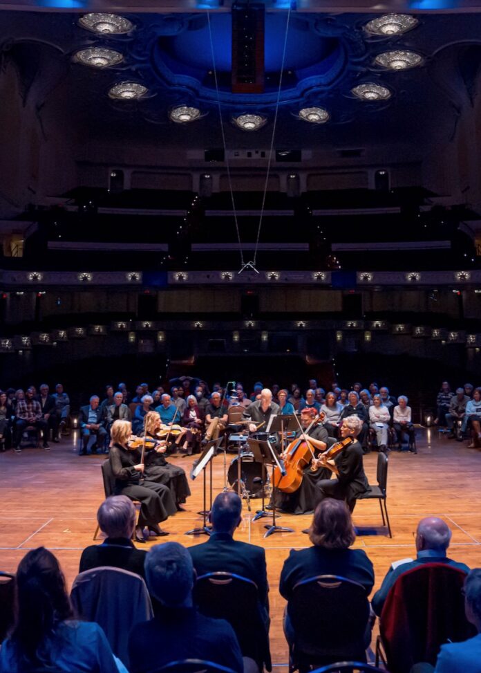 A small group of classical musicians play a concert onstage as people sit in a circle around them.