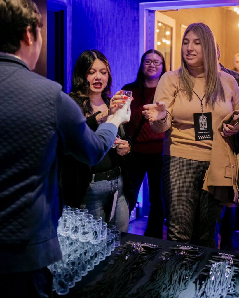 Women grab small glasses from a man at a table.