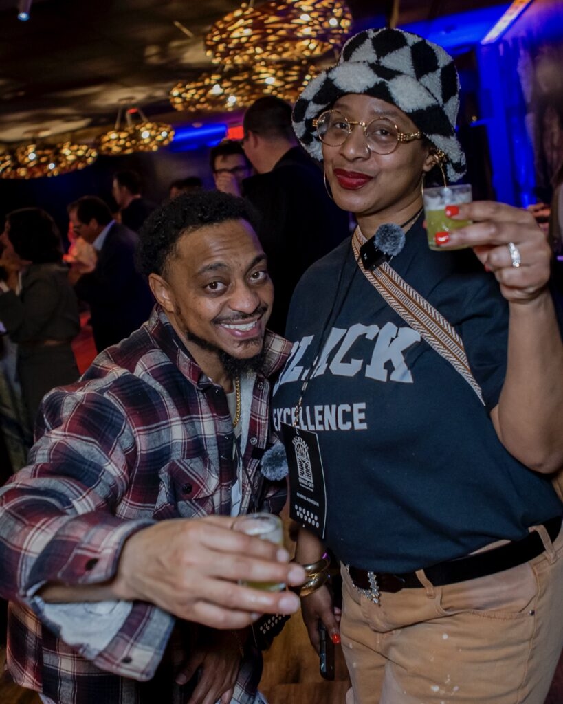 A woman holds a small cocktail glass with a man bending down beside her.