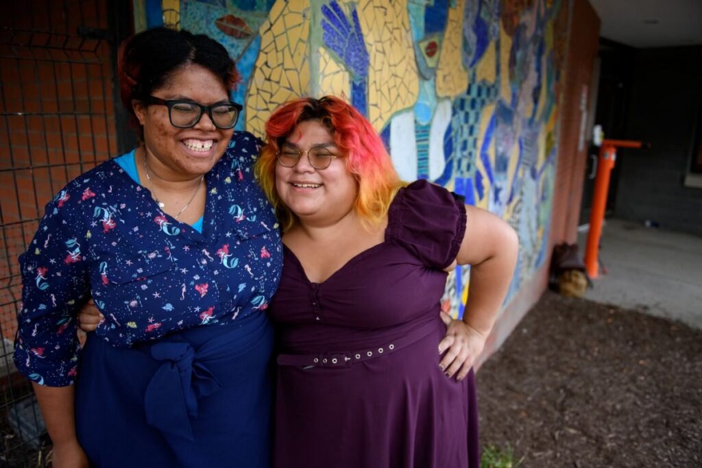Two smiling women standing together in front of a colorful mosaic mural as part of a community human services initiative.