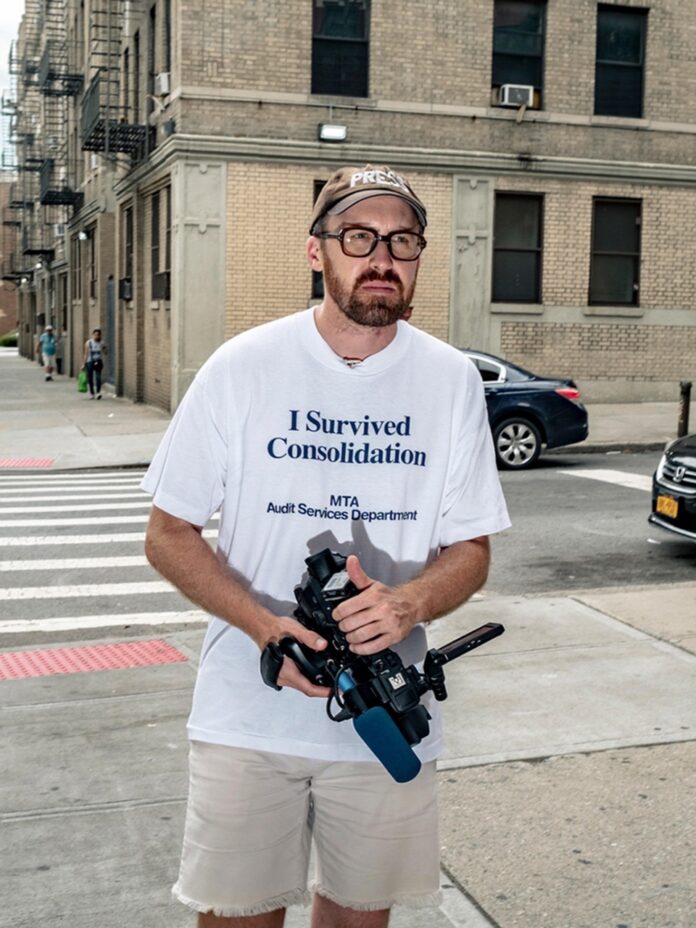 Conor Hannon stands in a white t shirt with a baseball cap on and a camera in his hands.