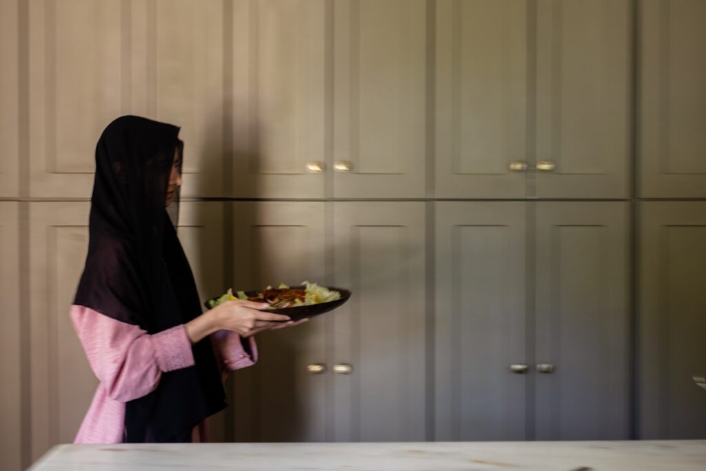 A woman in a black hijab and pink dress carries a finished plate of traditional Afghan food in a modern kitchen with minimalist cabinetry.