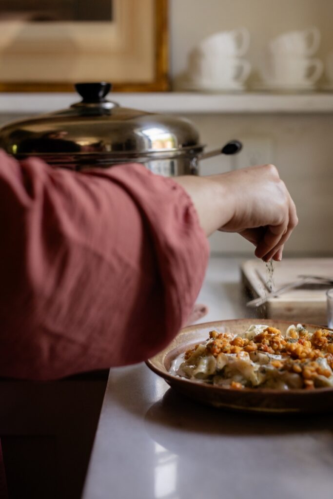 A close-up of Fatima’s hand garnishing a plate of Afghan mantoo dumplings with fresh coriander in a kitchen with a tiered steamer in the background.