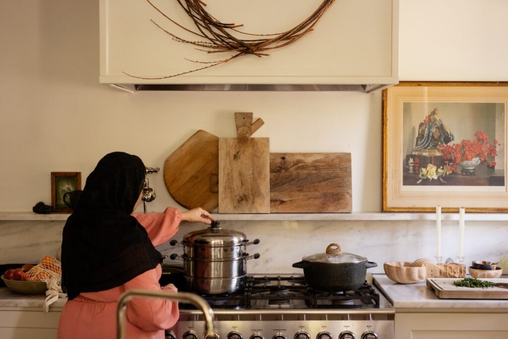 A woman in a hijab standing in a bright kitchen, carefully tending to a tiered metal steamer on a gas stove while preparing traditional Afghan mantoo.