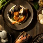 Filipino White Adobo Seafood Bouillabaisse featuring sautéed scallops, prawns, clams, and mussels on a bed of white garlic rice puree, garnished with fresh dill and fennel.