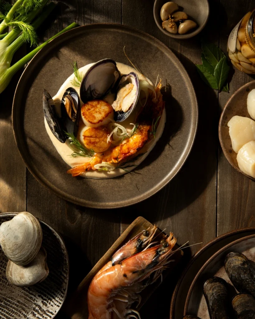 Filipino White Adobo Seafood Bouillabaisse featuring sautéed scallops, prawns, clams, and mussels on a bed of white garlic rice puree, garnished with fresh dill and fennel.