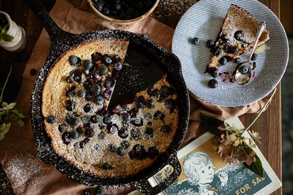 A dutch baby with blueberries sits in a cast iron pan with a slice on a plate next to it.
