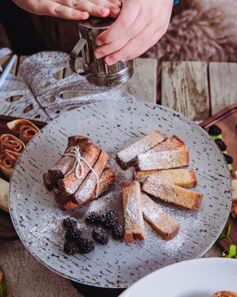 French toast sticks being dusted with powdered sugar, served with fresh blackberries on a decorative plate at a themed party.