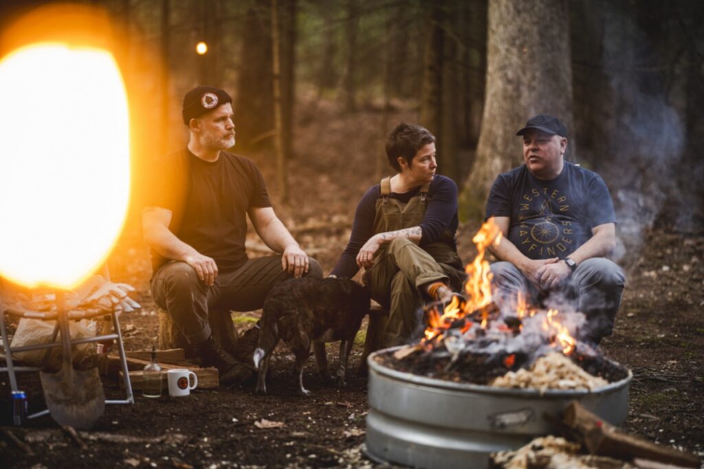 A group of friends sits around a roaring campfire in the woods with a dog, watching a primitive pottery firing in a galvanized metal pit.