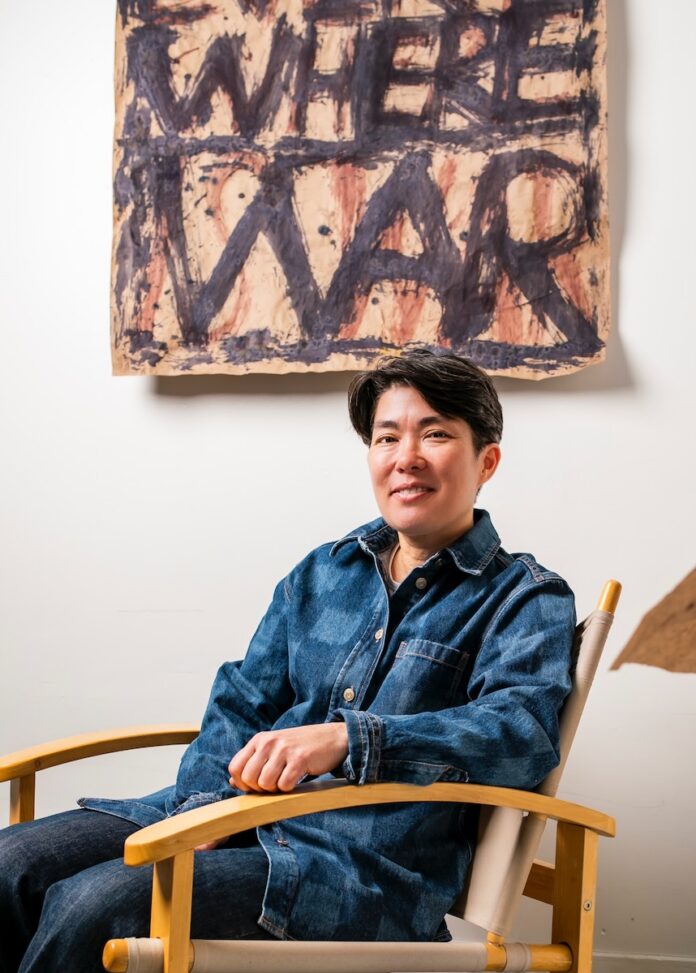 Ginger Brooks Takahashi sits in a wood chair in her studio with a painting above her.