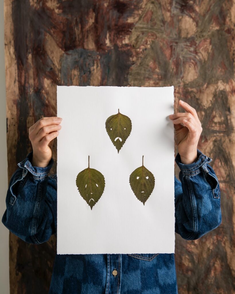 Hands hold up paintings of leaves with frowny faces.