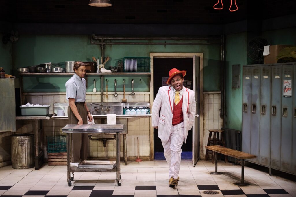 A man in a play walks on stage in a white suit and red hat while the other stands at a kitchen sink.