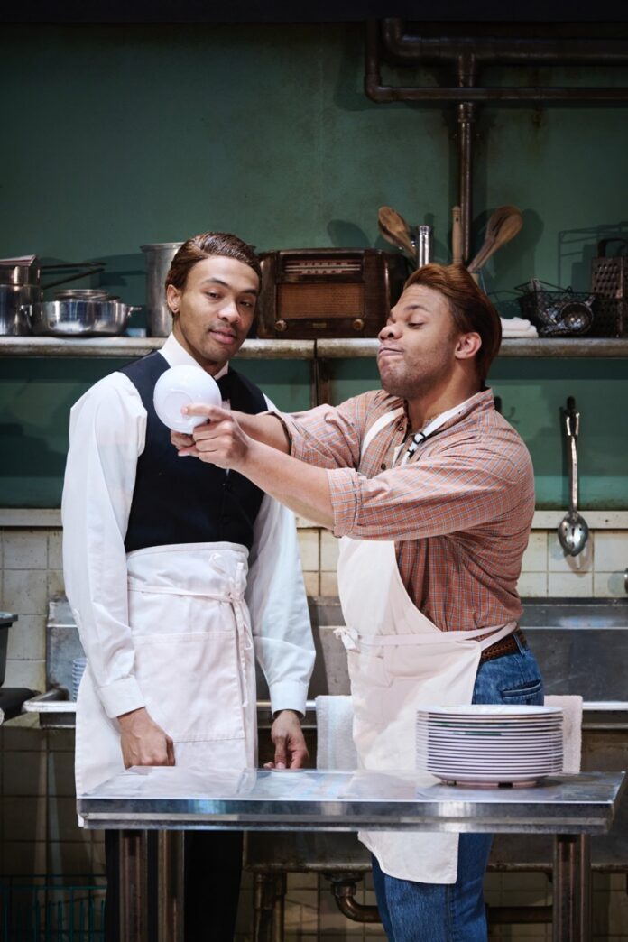 Two men on stage look at a bowl in a kitchen scene area.