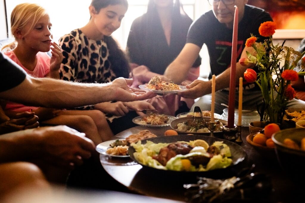 A diverse group of children and adults from two families sitting together in a dimly lit room, sharing a traditional Afghan feast featuring Kabuli Pulao and colorful flowers.