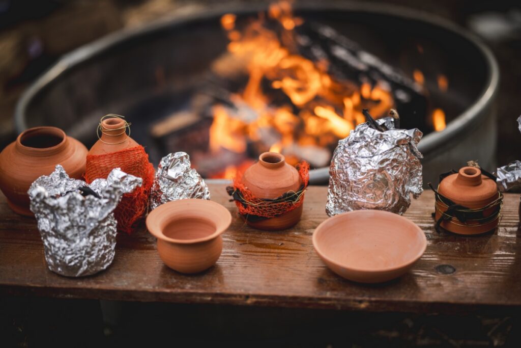 Handmade clay pottery pieces wrapped in copper wire and foil sitting on a wooden bench in front of a roaring campfire for primitive outdoor firing.