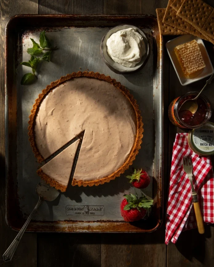 HoneyRum Strawberry Jam Semifreddo Tart in a graham cracker crust with a single slice removed, surrounded by fresh strawberries, whipped cream, and honeycomb.