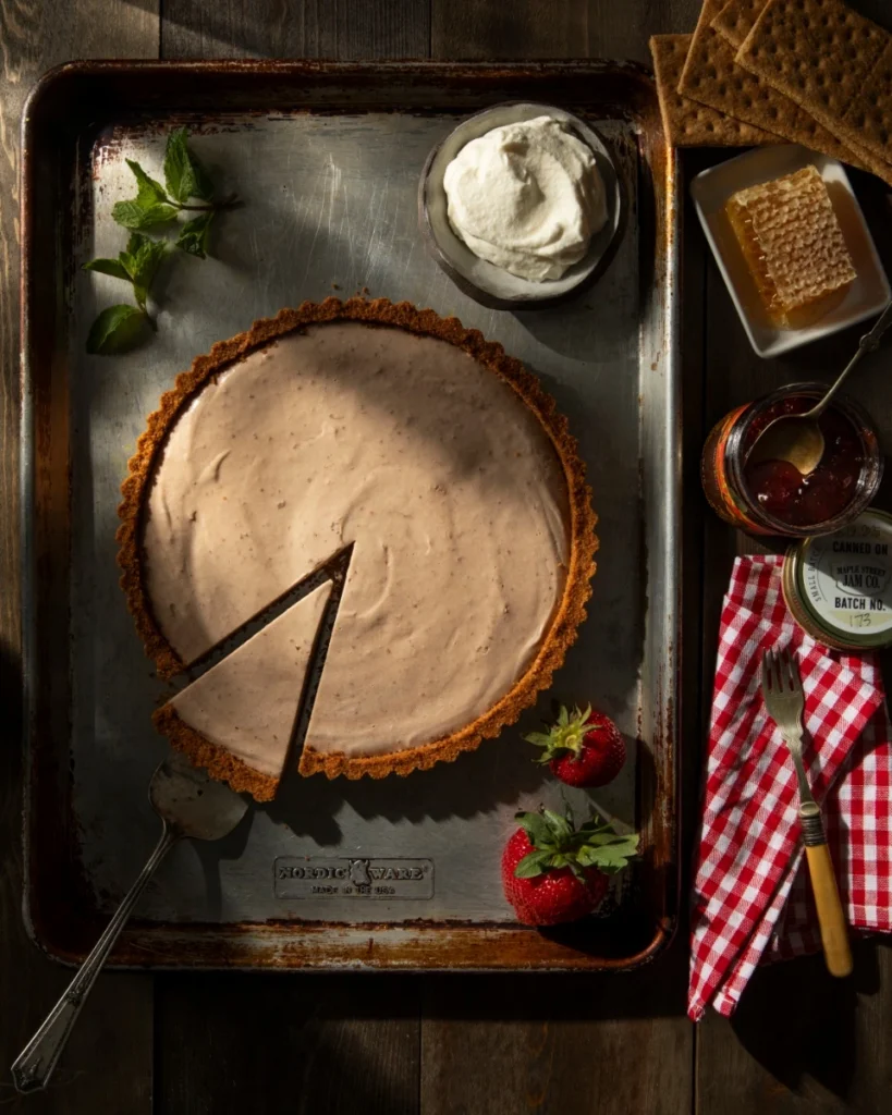 HoneyRum Strawberry Jam Semifreddo Tart in a graham cracker crust with a single slice removed, surrounded by fresh strawberries, whipped cream, and honeycomb.