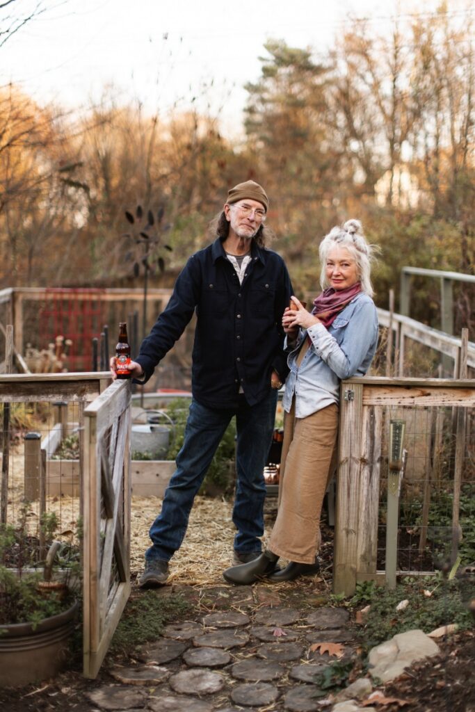 Hosts Cynthia and Mark standing by a wooden garden gate with seasonal beers and cider during an autumn outdoor gathering near Pittsburgh.