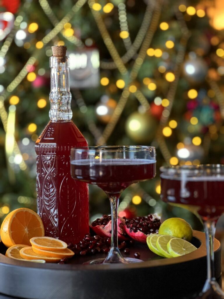 A fancy bottle with a deep red Good Luck Sparkling Pomegranate Cosmo on a tray with 2 cocktail glasses, orange slices, an open pomegranate, and lemon slices in front of a blurred Christmas tree.