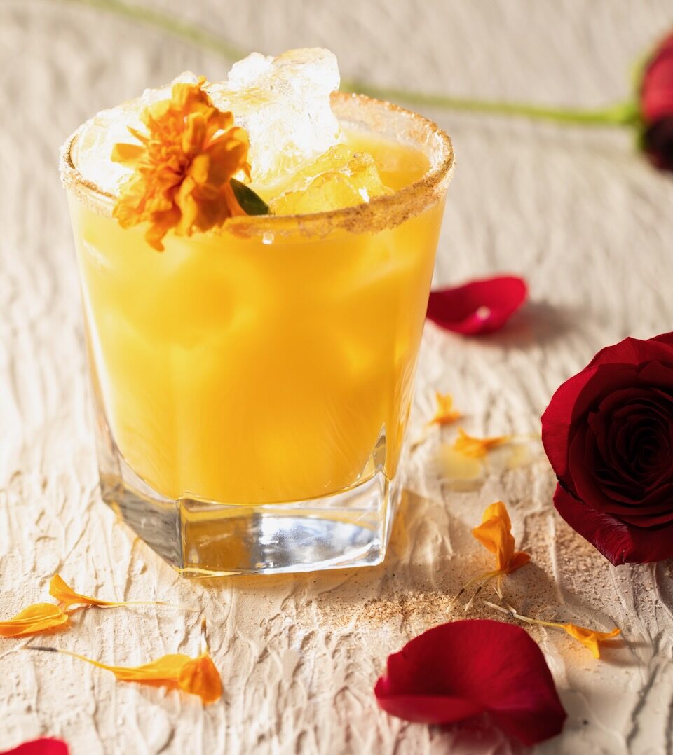 A short cocktail glass has an orange cocktail inspired by 10 Things I Hate About You with a cinnamon sugar rim and edible marigold inside. On the table around the cocktail lays marigold petals and red rose petals.