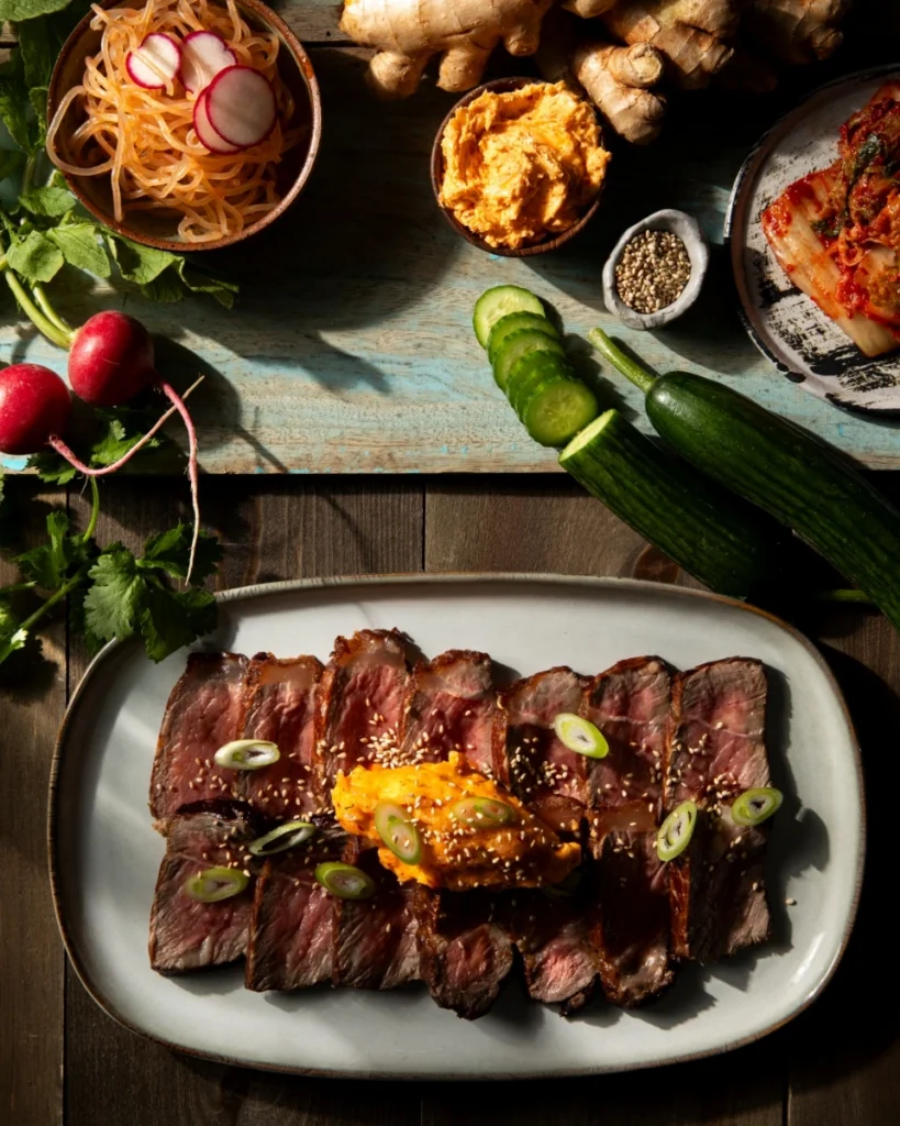 Sliced steak topped with a scoop of orange kimchi butter and garnished with scallions and sesame seeds, surrounded by fresh ingredients including ginger, sliced cucumbers, and radishes.