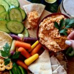 A bowl full of rotisserie buffalo chicken dip with pita bread, carrots, cucumbers, chips, and pretzels around it.