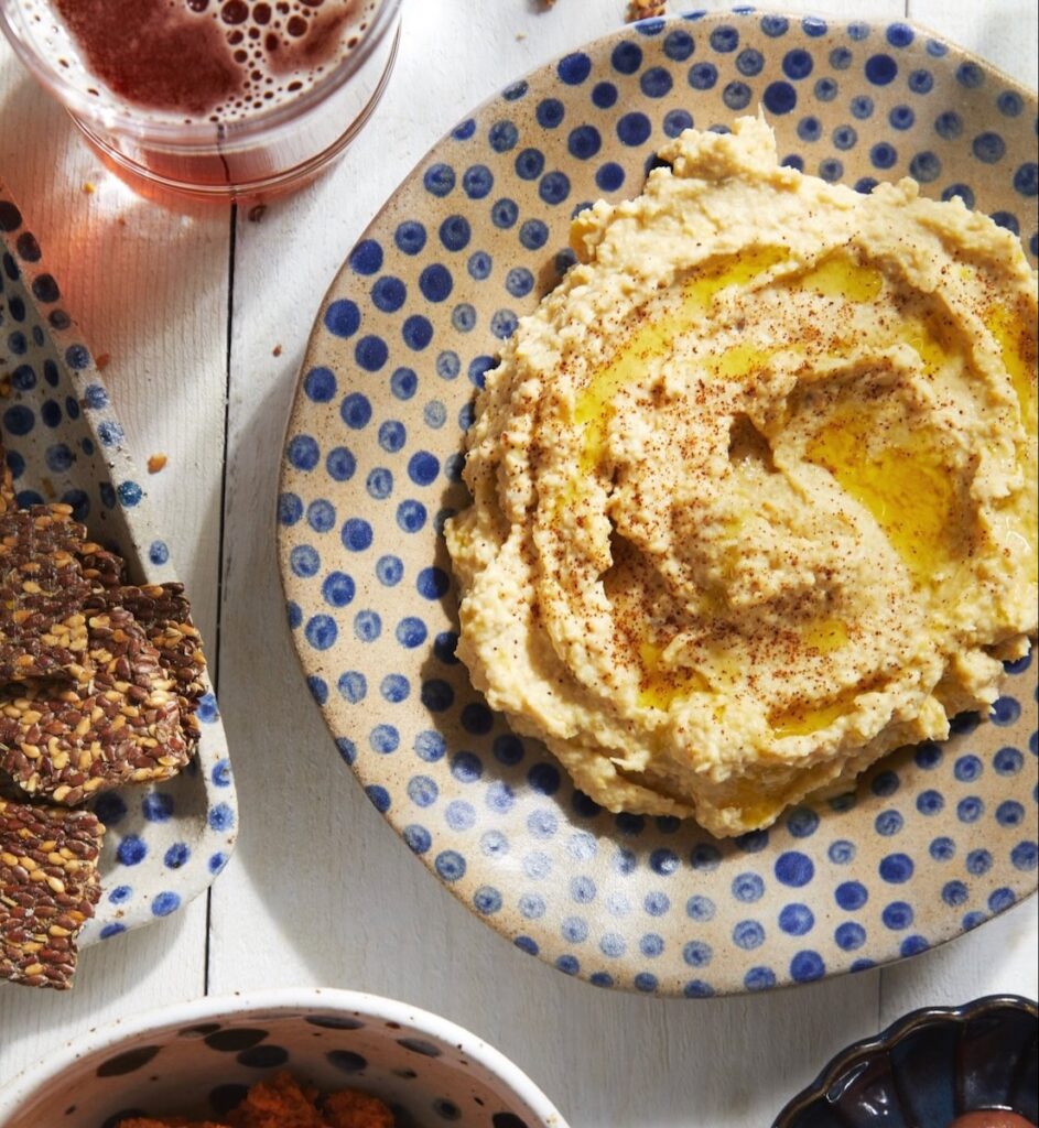 A lemon artichoke hummus sits in a polka dot blue and white bowl on a white picnic table.