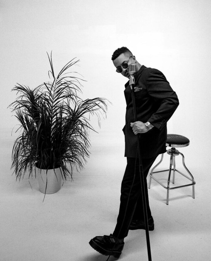 A man stands in front of a stool and plant holding a mic stand.