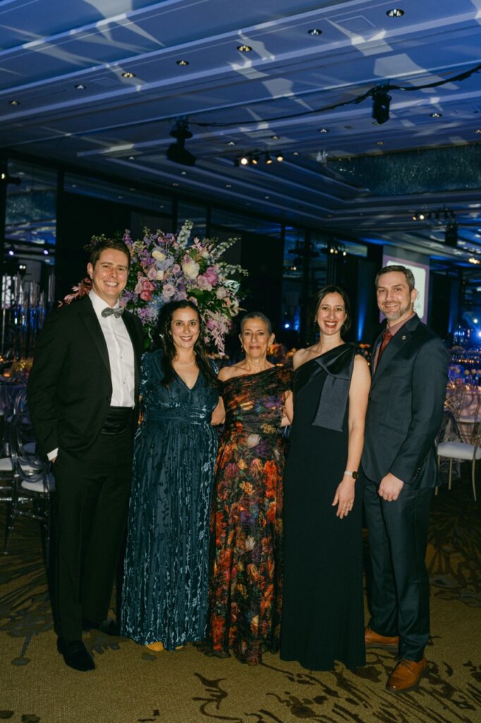 A group of women in dresses and men in tuxes stand together in blue lights.