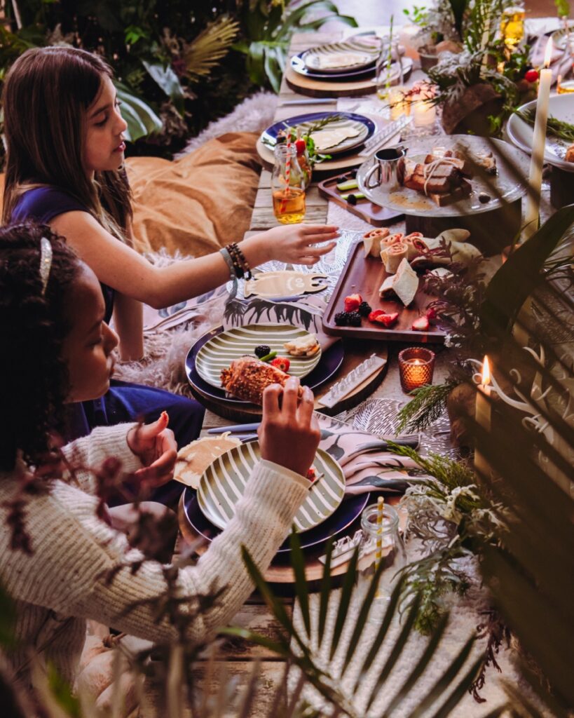 Two young girls sitting on floor cushions at a "Where the Wild Things Are" party table, enjoying cheeseburger croissants and sun butter pinwheels.