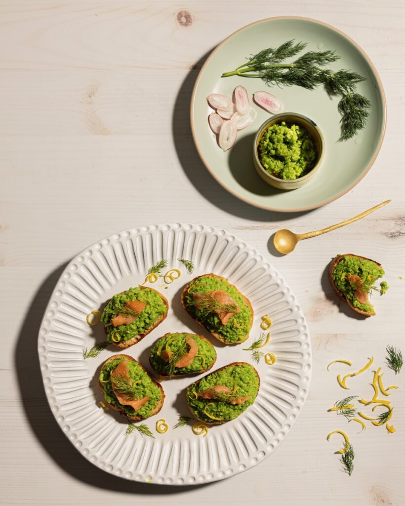 A white plate holds a pea puree bruschetta with smoked salmon on top as a green plate off to the side holds a small bowl of the pea purree.
