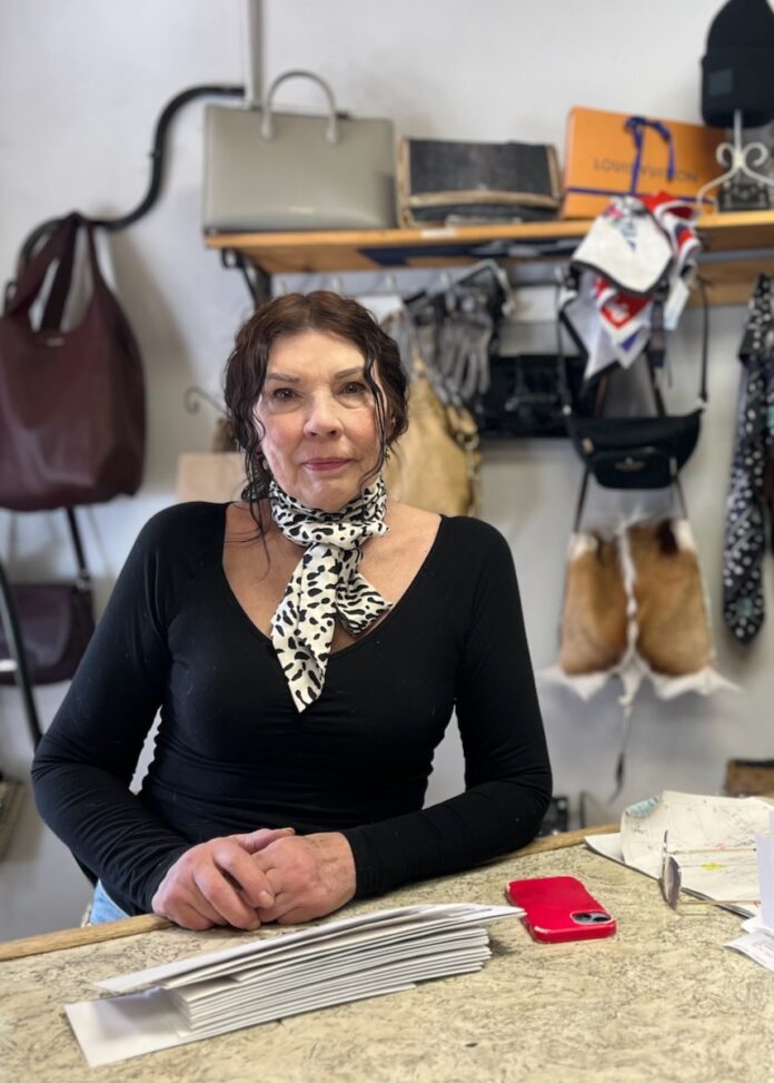 Jacqueline Stone stands at a table with purses on a shelf behind her.