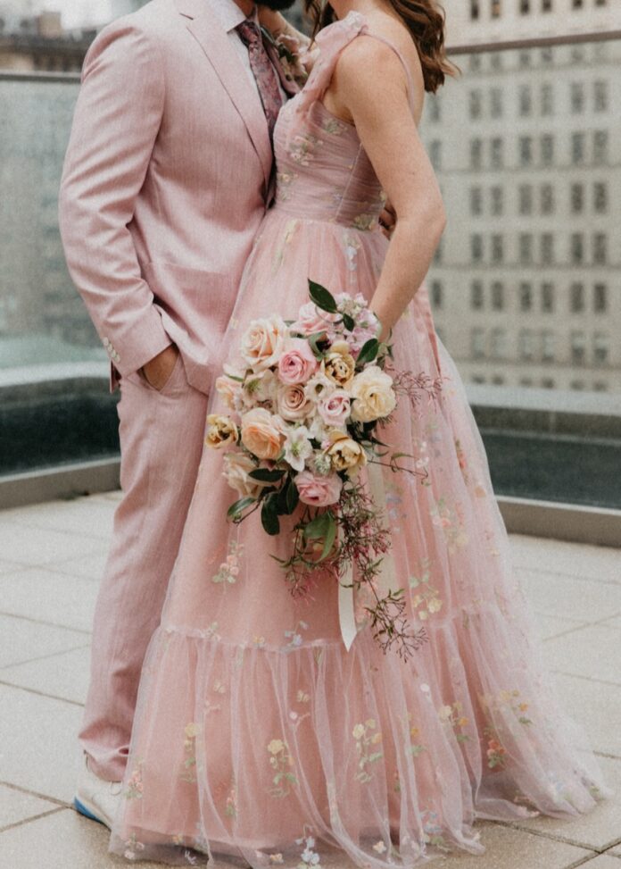 A man in a pink suit and a woman in a long pink dress stand on a roof holding a bouquet of flowers.