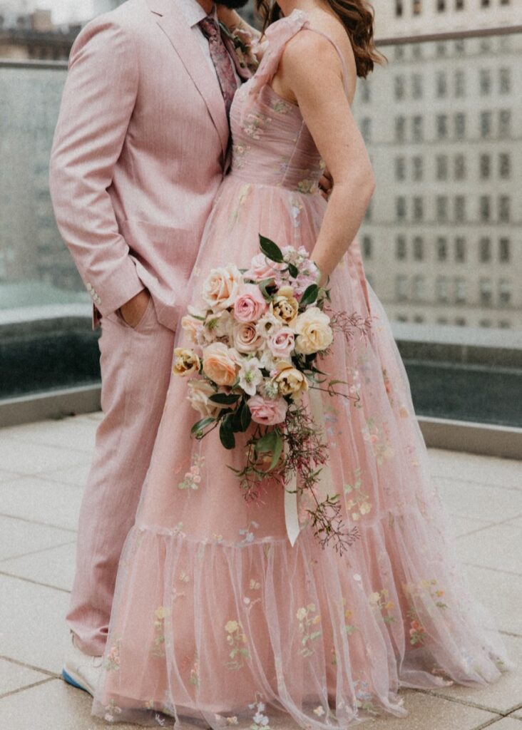 A man in a pink suit and a woman in a long pink dress stand on a roof holding a bouquet of flowers.