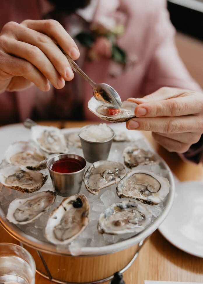 A man puts a red sauce onto a pail full of oysters at a table.
