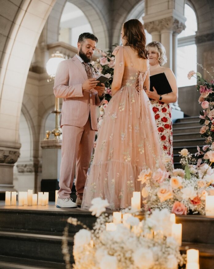 A man and woman hold hands at a wedding alter with flowers and candles surrounding them on the stairs.